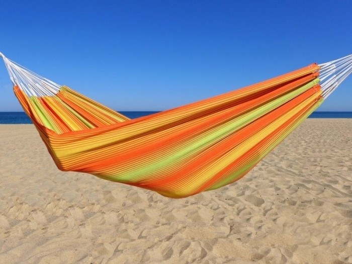 Hamac coloré en coton aux rayures orange, jaunes et vertes, installé sur une plage ensoleillée avec ciel bleu éclatant