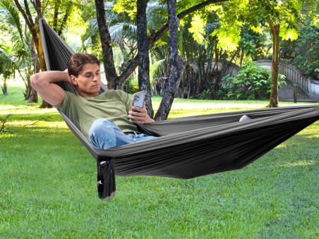Un garçon se repose et consulte son téléphone dans un hamac outdoor flex XL noir et gris au milieu d'une forêt
